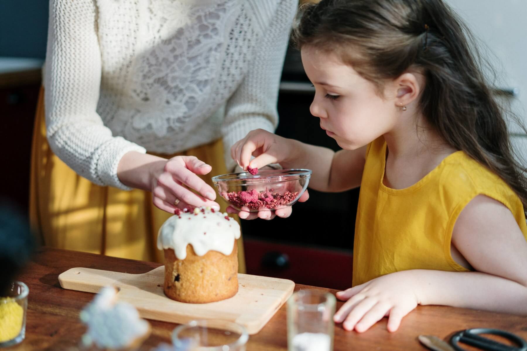 Young girl sprinkling toppings on a dessert with her mother