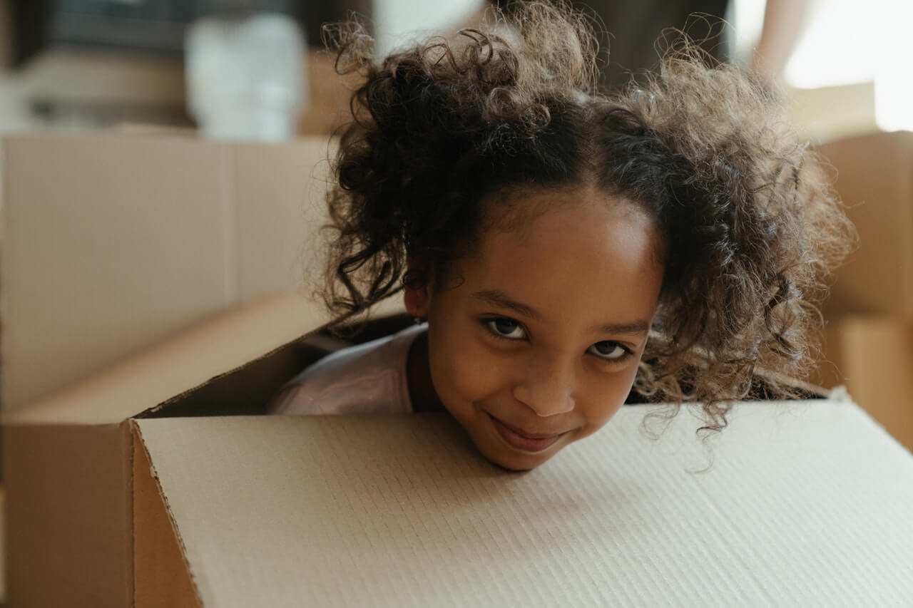 Young girl peeking out of a large cardboard box. Photo credit: Cottonbro