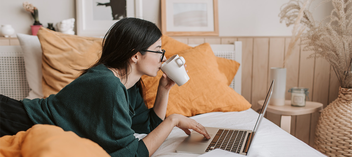 Woman lying in bed, drinking coffee, and working on a laptop. Image credit: Vlada Karpovich